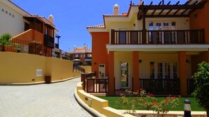 Un edificio residencial con una entrada de vehículos curvada, un balcón y un pequeño jardín con flores rojas.