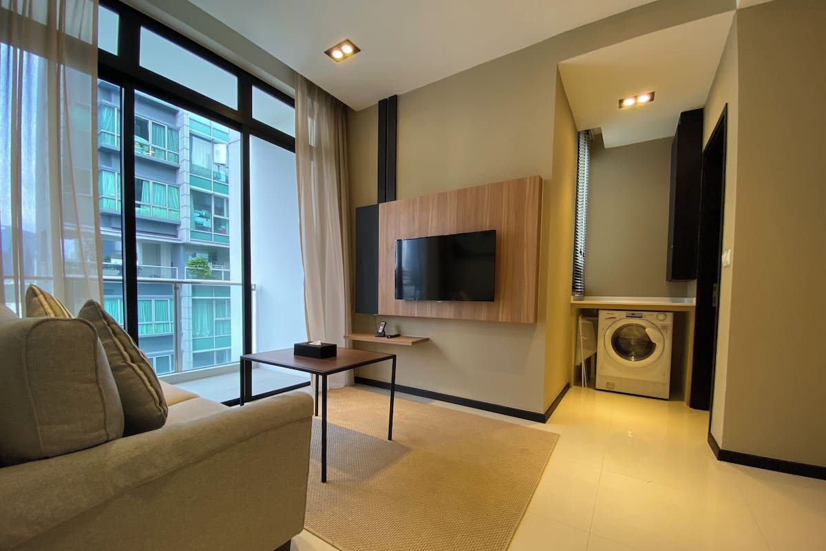 Ein modernes Wohnzimmer mit einer Couch, einem kleinen Tisch, einem an der Wand befestigten Fernseher und einer Waschmaschine in der Ecke.