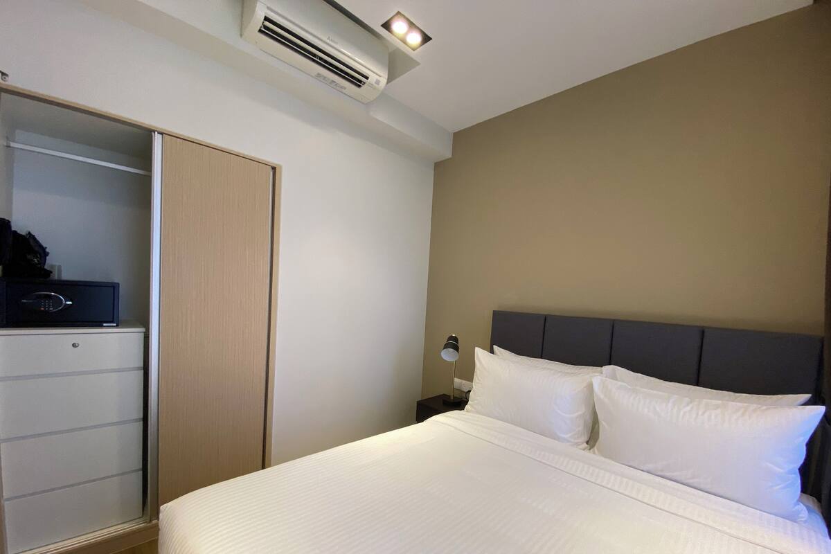 A hotel room with a bed, a wardrobe, a bedside table with a lamp, and an air conditioning unit.