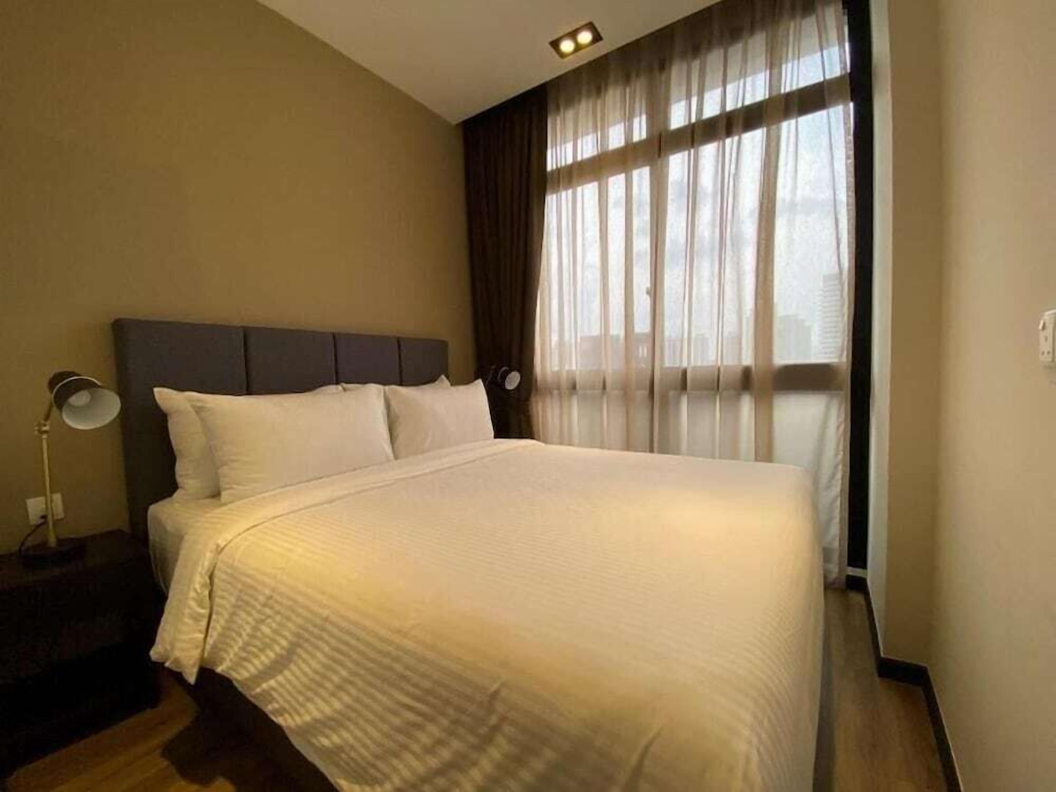 A neatly made bed with a headboard, a bedside lamp, and a window with sheer curtains.