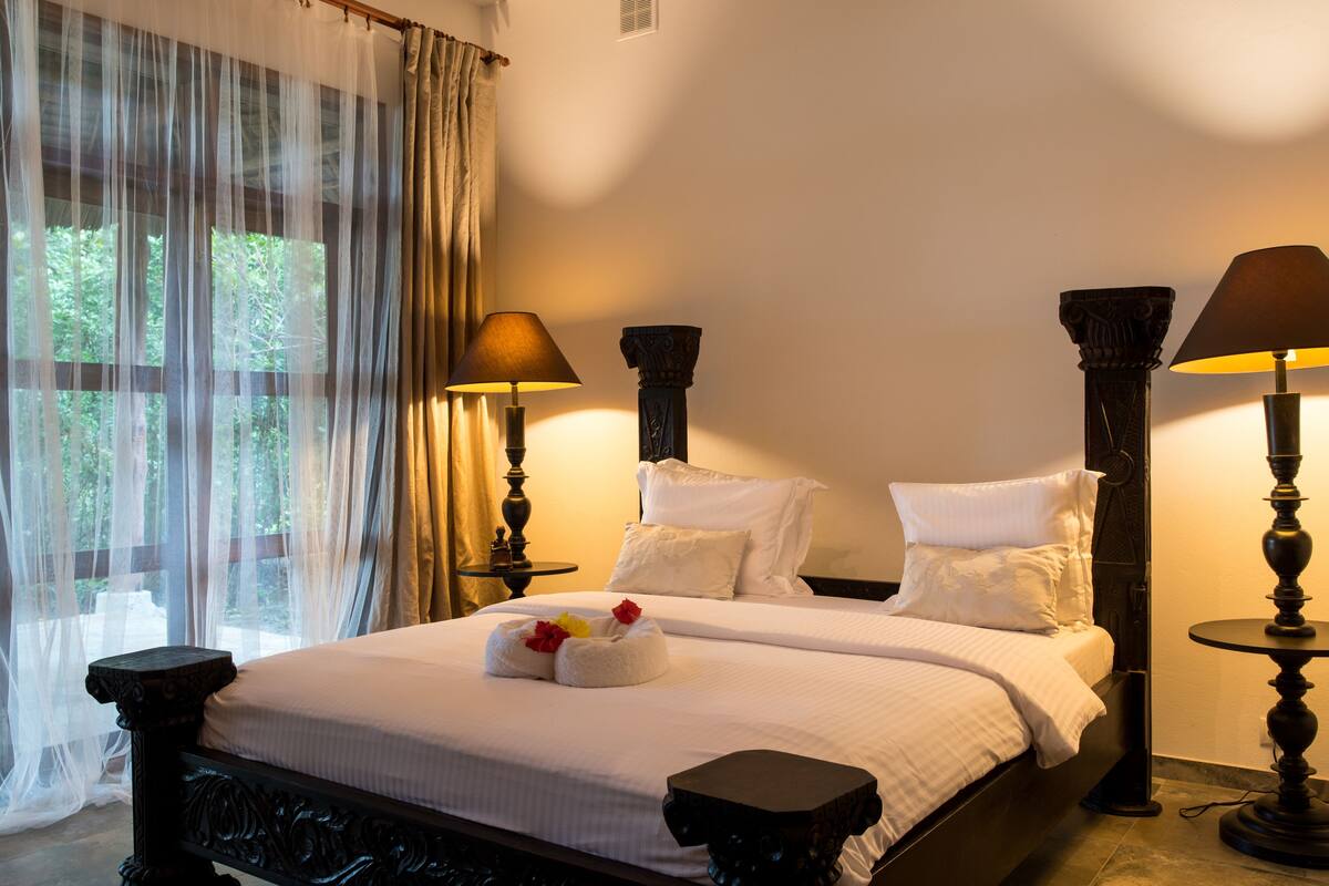 Cama doble con ropa de cama blanca, dos mesitas de noche con lámparas, cortinas translúcidas y vistas al exterior.