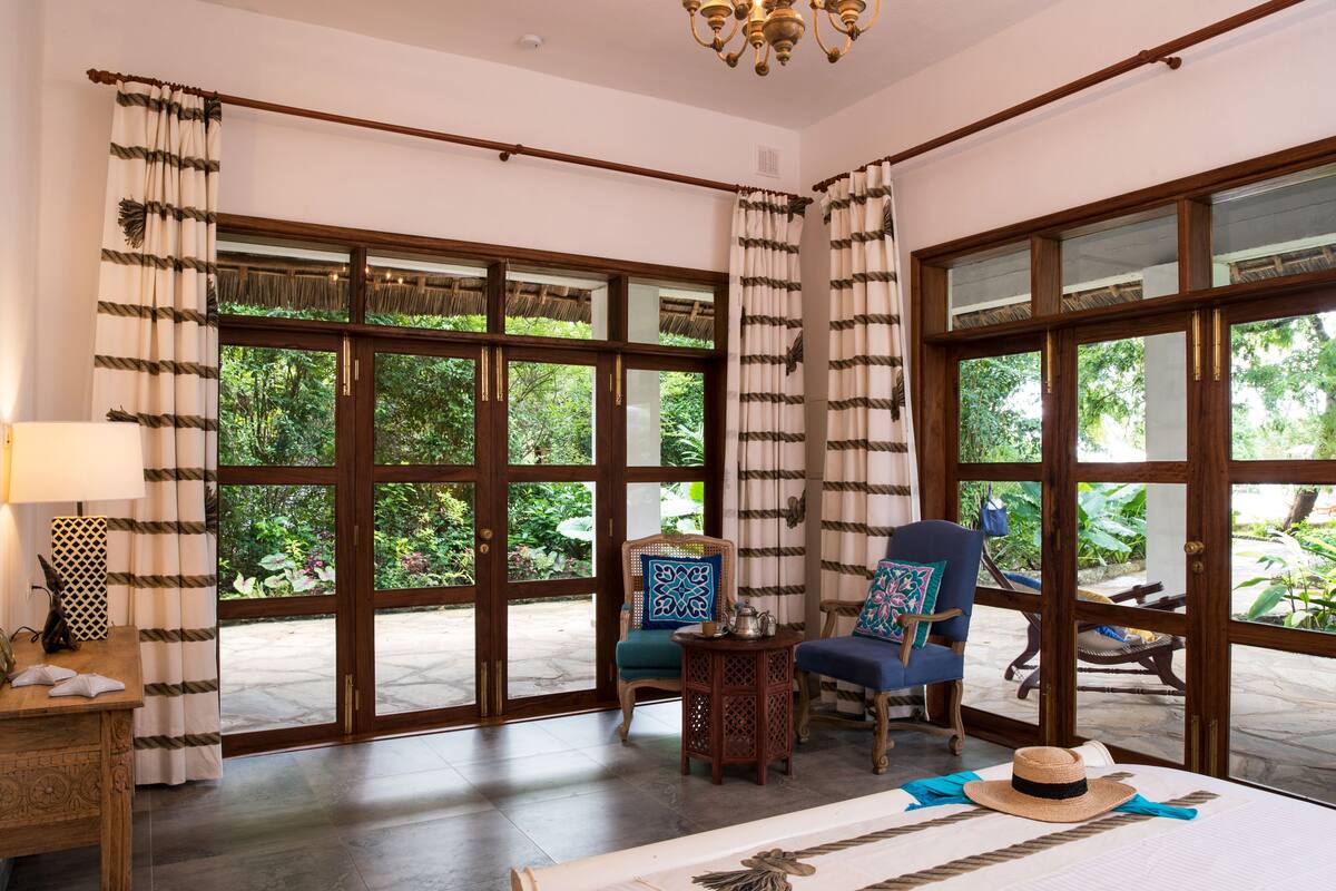 Una habitación con ventanas amplias, una mesa de madera, dos sillas y una mesita con una lámpara.