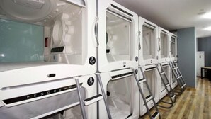 Een rij witte, moderne, gesloten massage stoelen met metalen frames en bedieningspanelen, opgesteld langs een lichtgekleurde muur in een ruimte met een houten vloer.