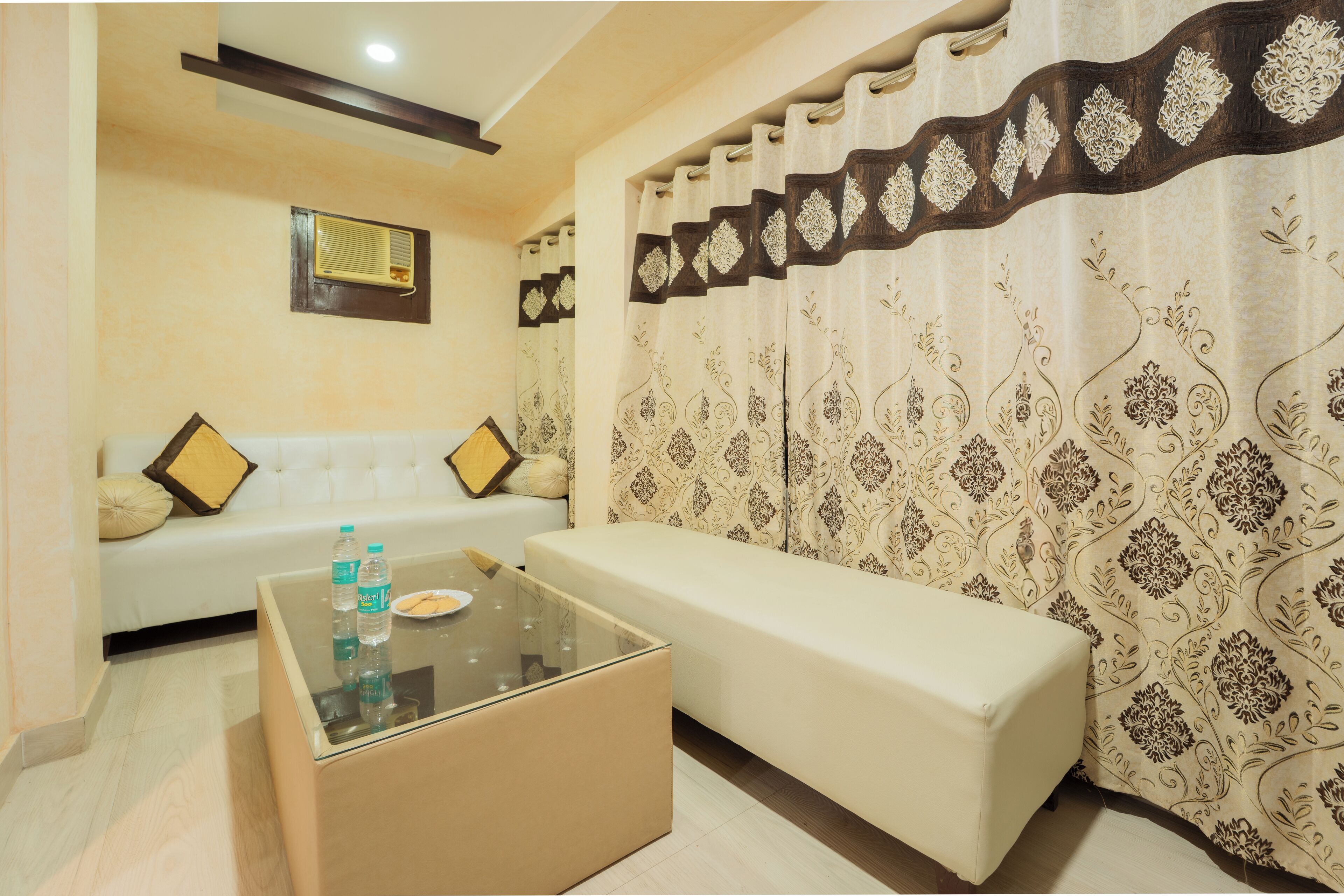 A room with a sofa, a bench, a glass coffee table, and patterned curtains.