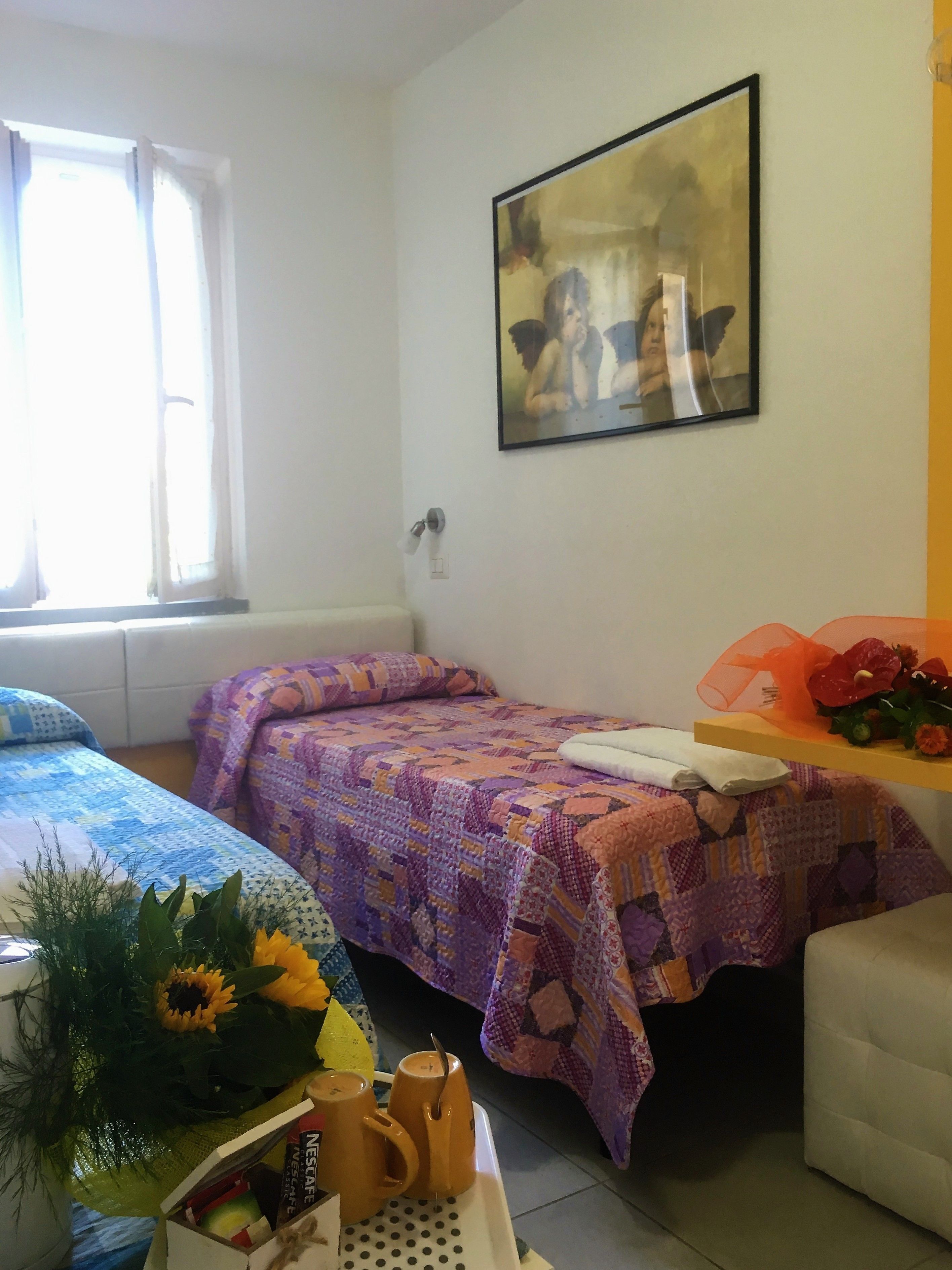 A room with two beds, a window, a painting, and a table with a bouquet and coffee mugs.