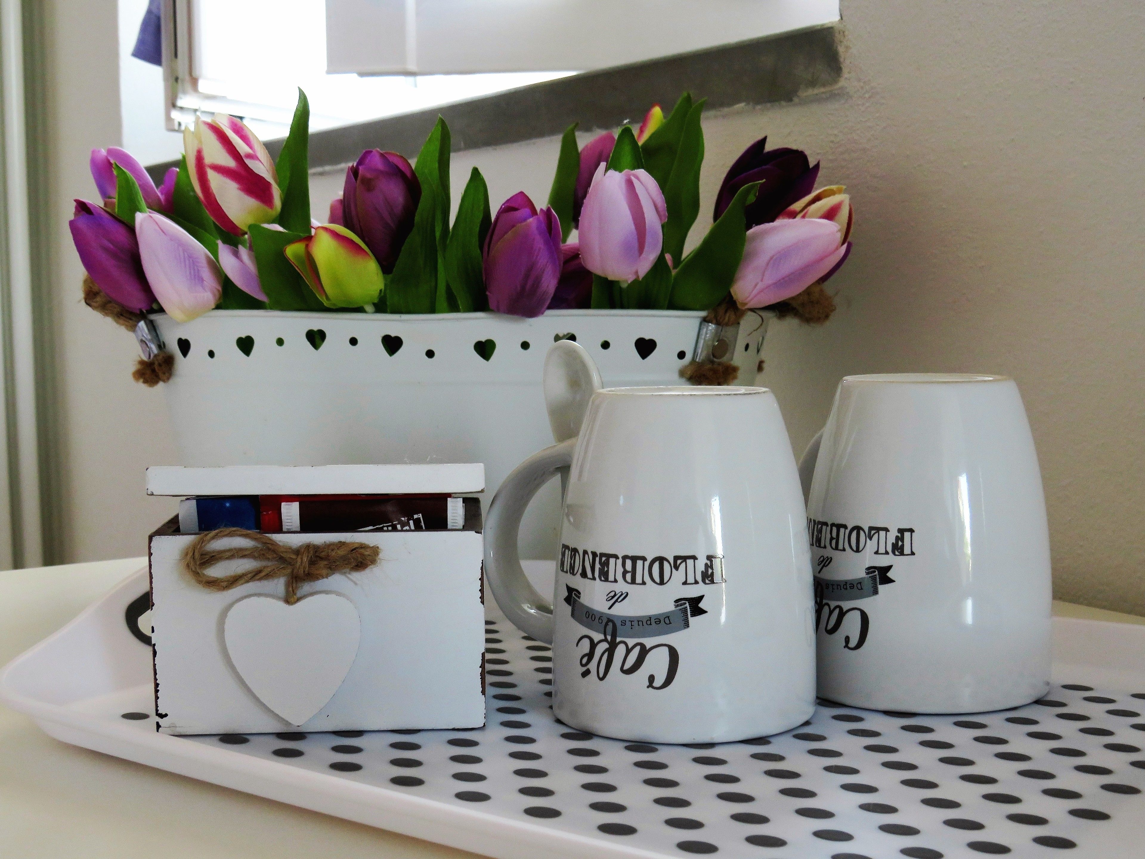 Ein Tablett mit einer weißen Tasse, einer kleinen weißen Schachtel mit einem Herz und einer Vase mit violett-rosa Tulpen.
