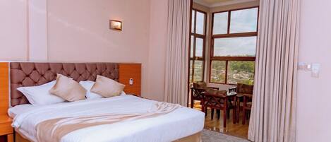 A hotel room with a large bed, a headboard, a small table with chairs, and a view through a large window.