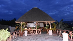 A thatched-roof structure with wooden furniture and potted plants, set against a night sky.