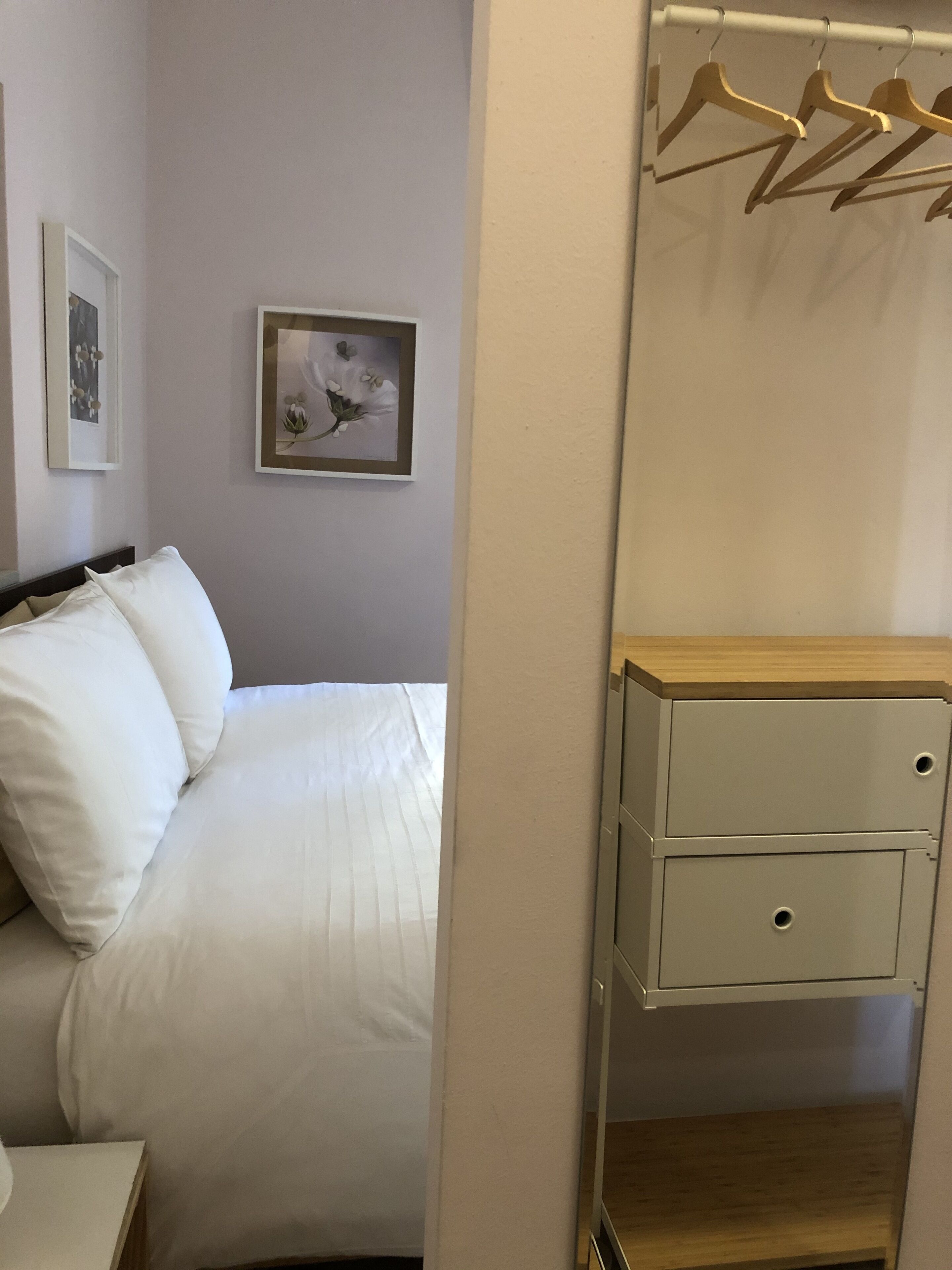 Ein ordentlich bezogenes Bett mit weißen Leinentüchern, ein kleiner Nachttisch und ein Nachttisch mit Schubladen aus Holz. An der Wand über dem Bett befinden sich zwei gerahmte Bilder.