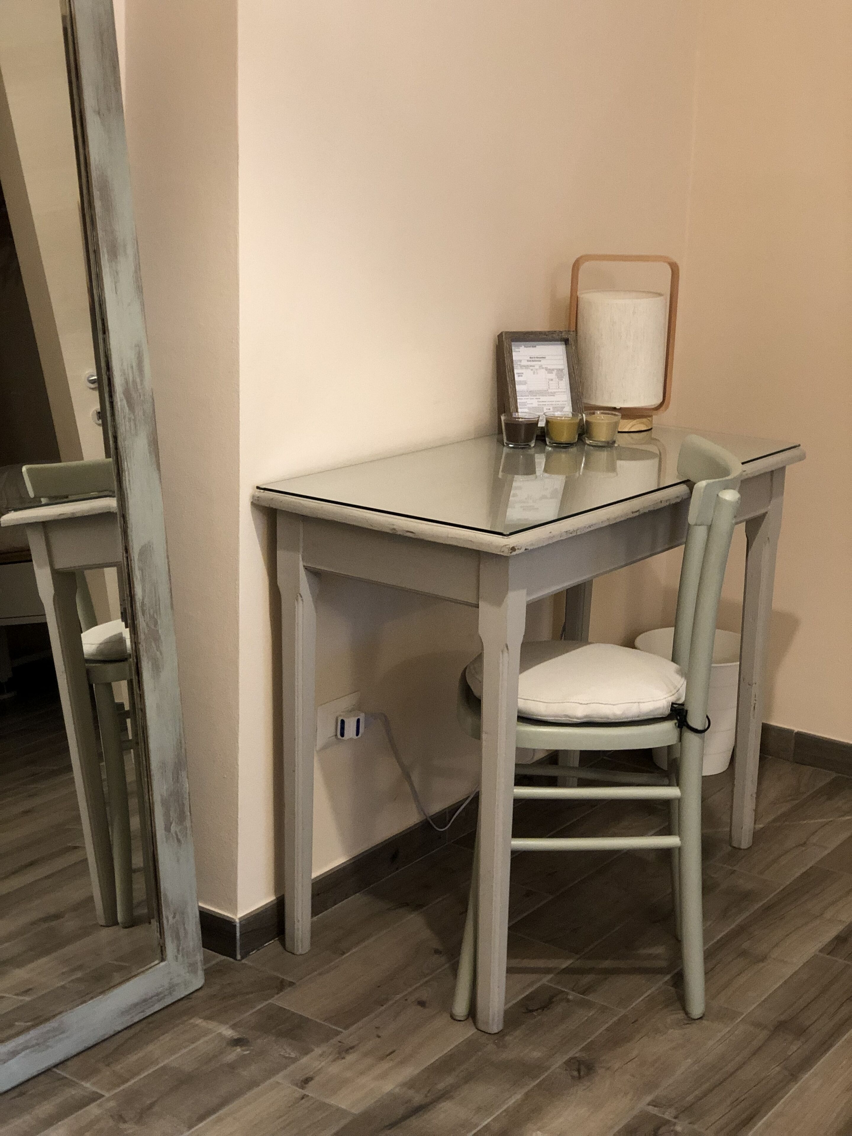 A small, glass-top desk with a chair, a lamp, and a menu stand in a corner.