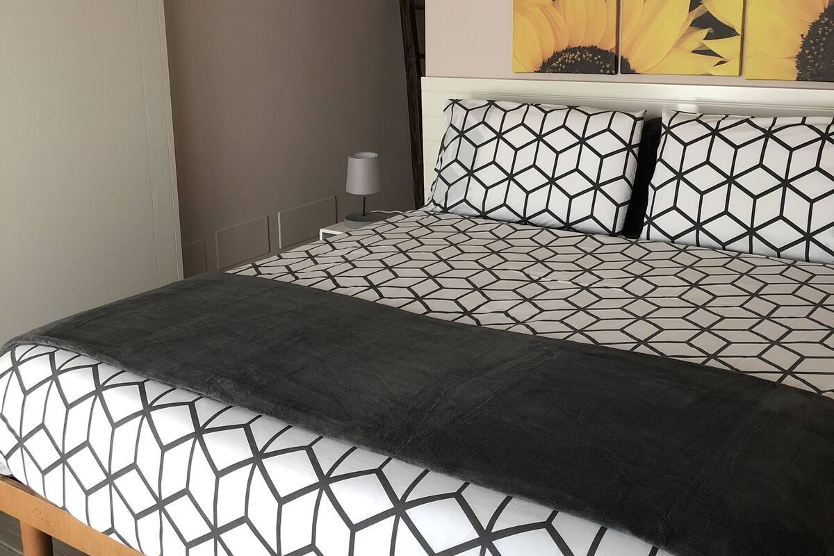A bedroom with a bed featuring a geometric patterned bedspread, a black throw blanket, and a wall-mounted air conditioning unit above the bed.