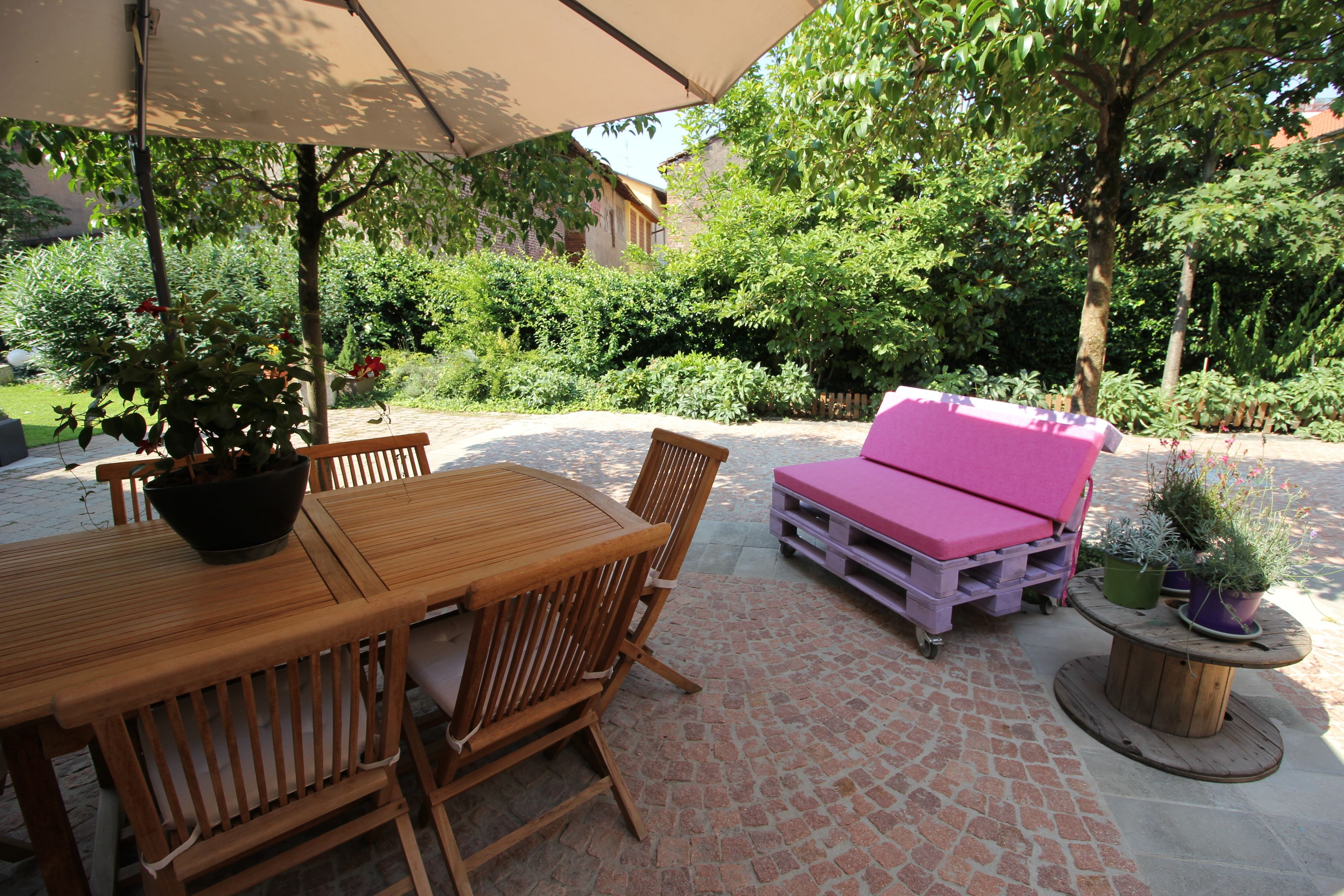 Un patio exterior con una mesa y sillas de madera, un sofá rosa y plantas en macetas.
