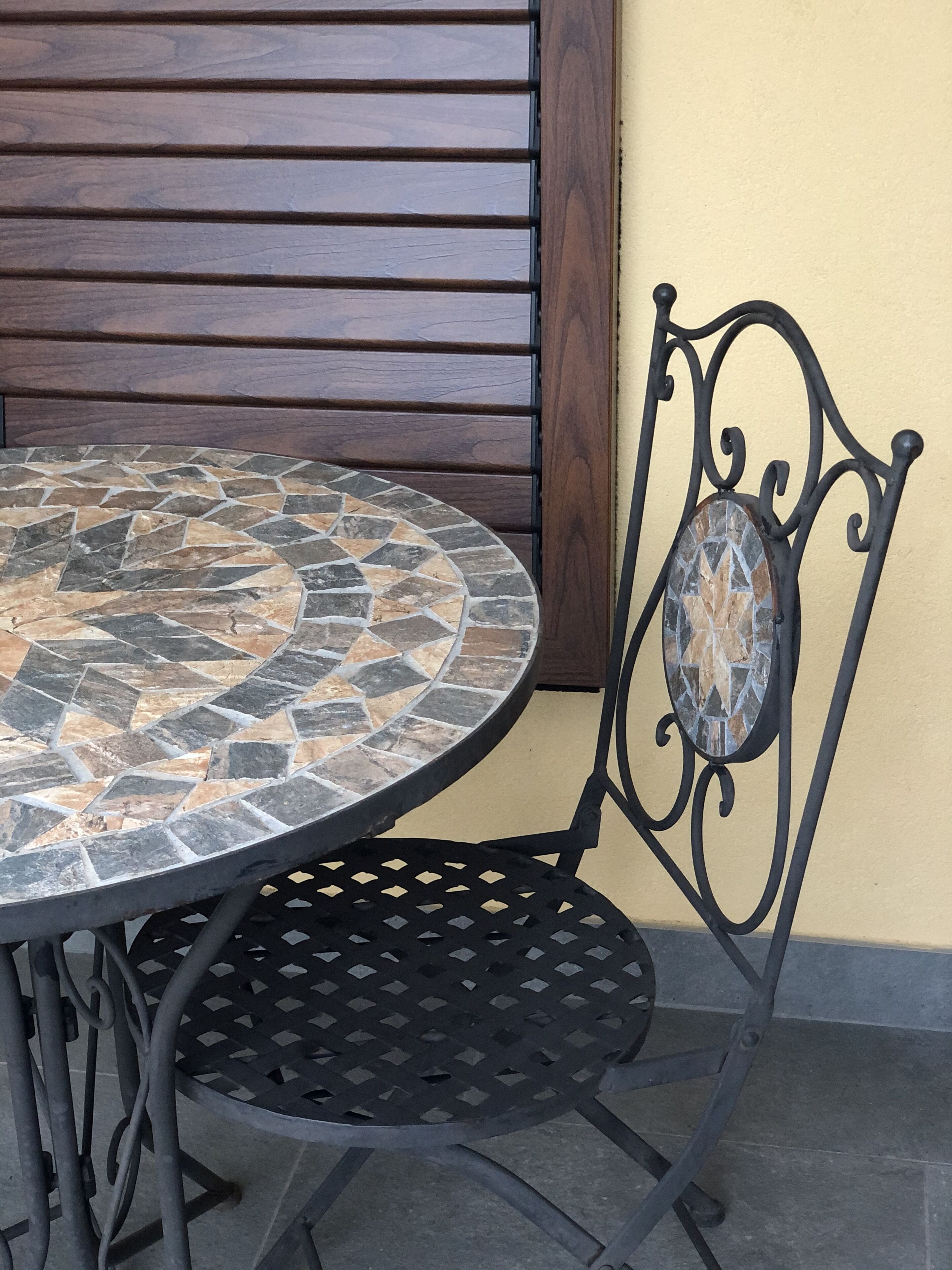 Una mesa redonda con una superficie de mosaicos, una silla de metal negro con un respaldo decorativo, y una pared de listones de madera al fondo.