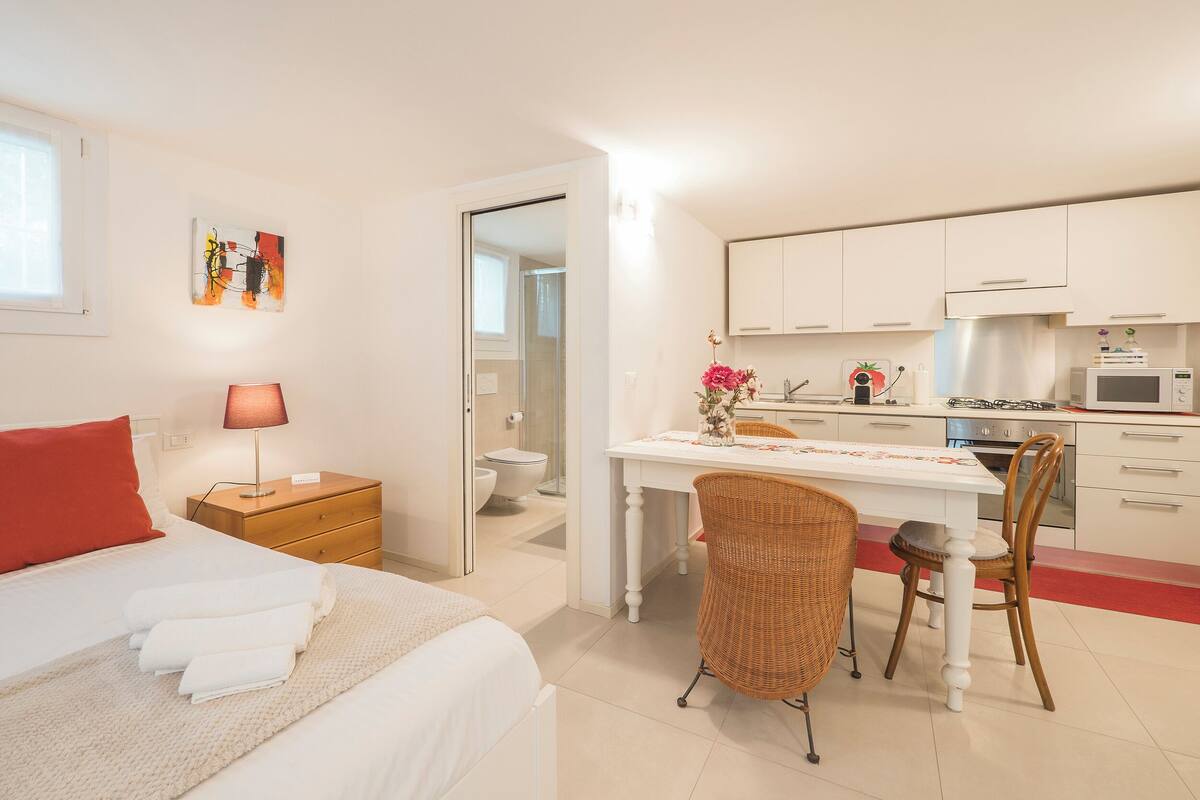 A compact hotel room with a bed, bedside table, lamp, and a view of the adjacent kitchen.