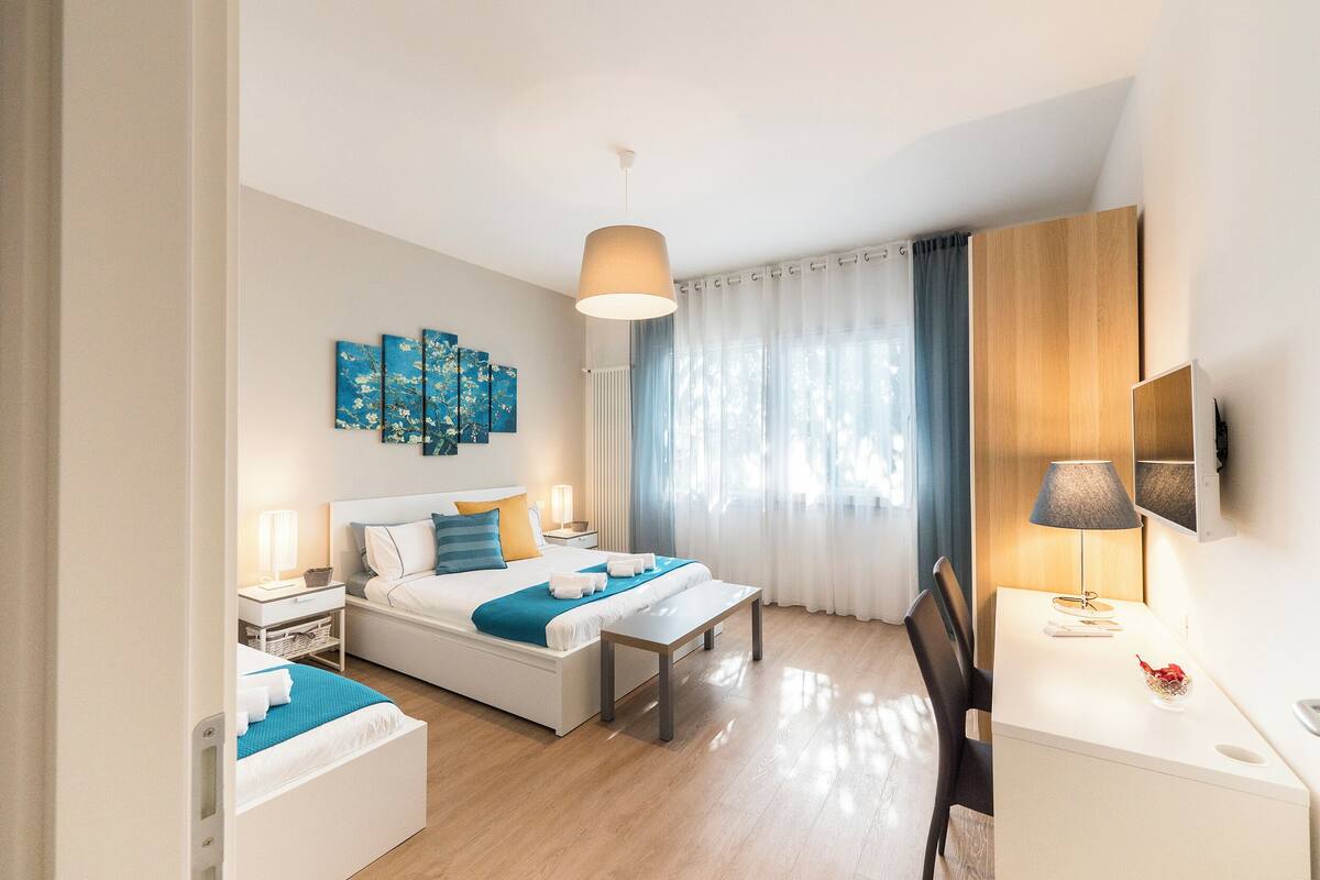 A modern hotel room with a bed, bedside tables, a desk, and a large window with curtains.