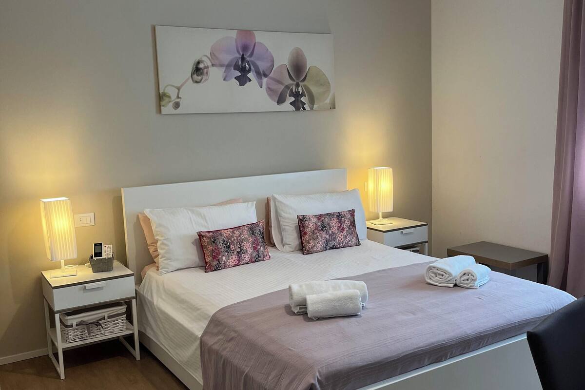 A neatly arranged bedroom with a large bed, two bedside tables with lamps, a wall-mounted artwork, and a wooden floor.