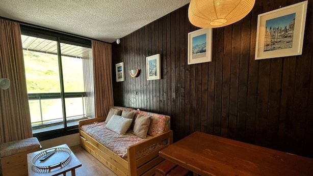 Una habitación acogedora con un sofá y una mesa de madera, además de un banco. Se observan cuadros en la pared y una ventana con cortinas.
