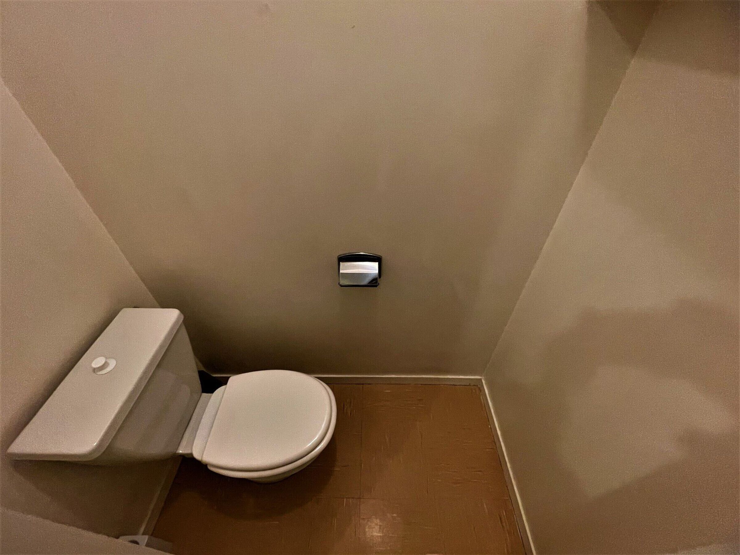A small, narrow bathroom with a toilet, a wall-mounted flush handle, and a wooden floor.