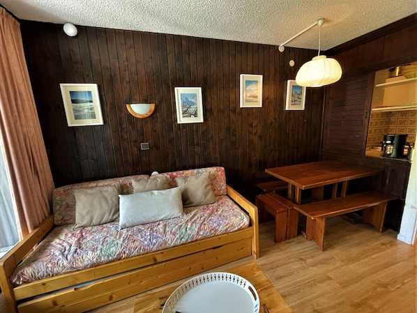 A cozy room with a wooden sofa, a wooden table, and a bench. There are framed pictures on the wall and a hanging light fixture.