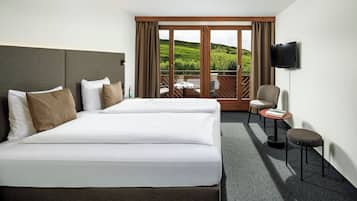 A hotel room with two beds, a TV, a small table with chairs, and a balcony with a view of a green landscape.