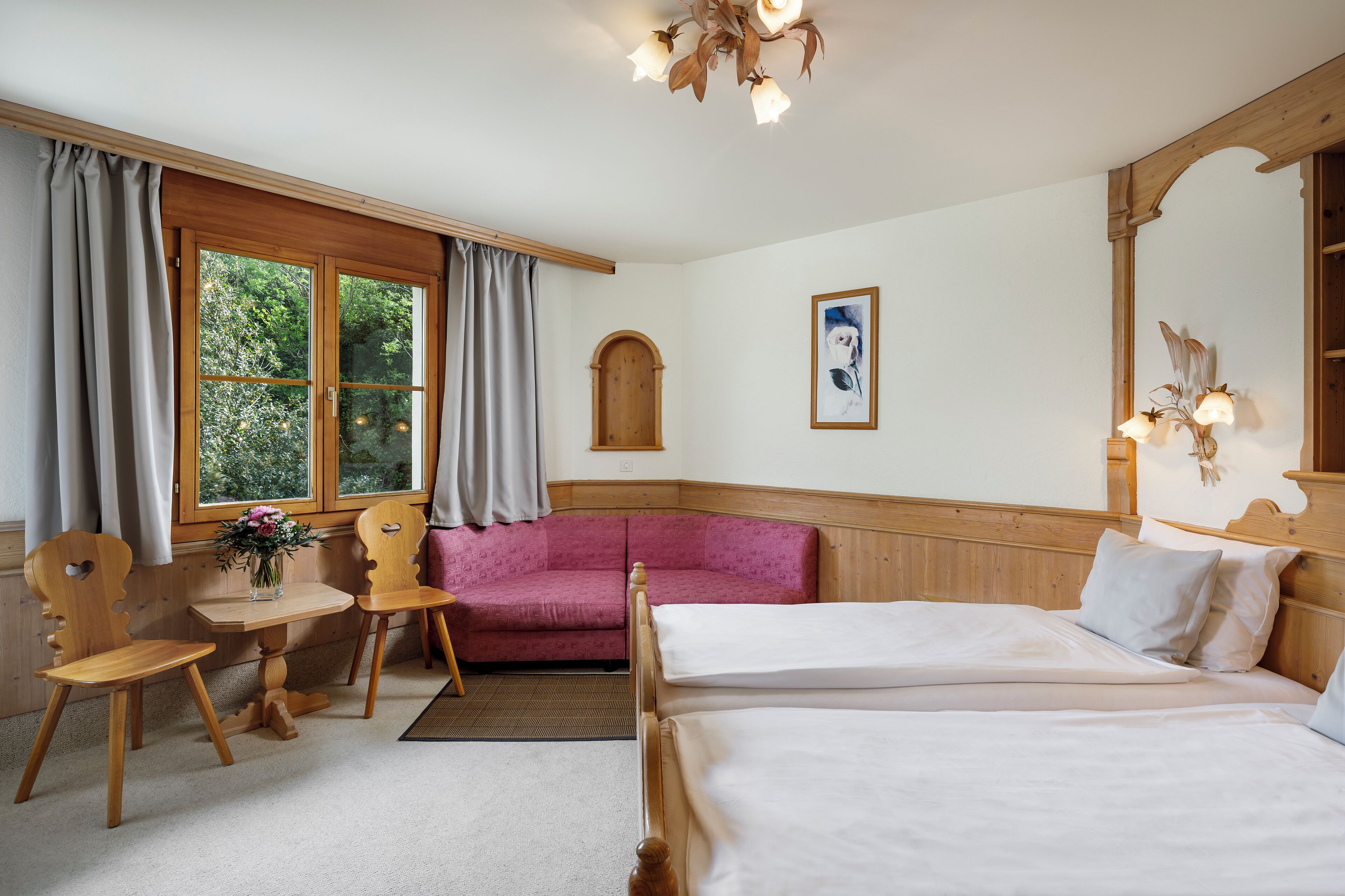 A hotel room with two beds, a wooden headboard, a pink sofa, a wooden table with a vase of flowers, and a window with curtains.