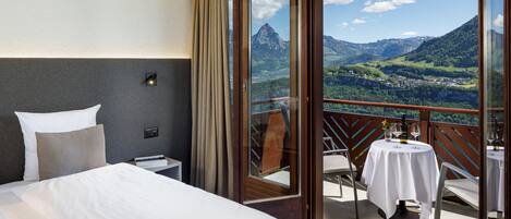 A hotel room with a bed, a nightstand, and a table with a tablecloth. There is a balcony with a table and chairs, and a view of mountains and a valley.