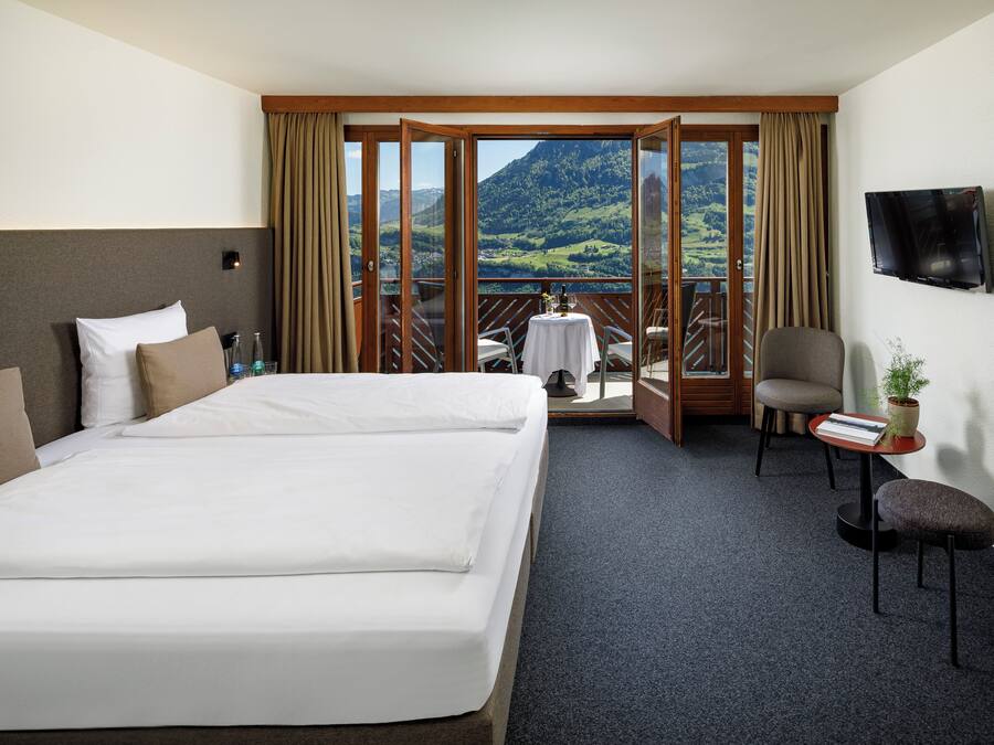 Habitación de hotel con una cama grande, una mesita con una planta, una silla y un televisor en la pared.