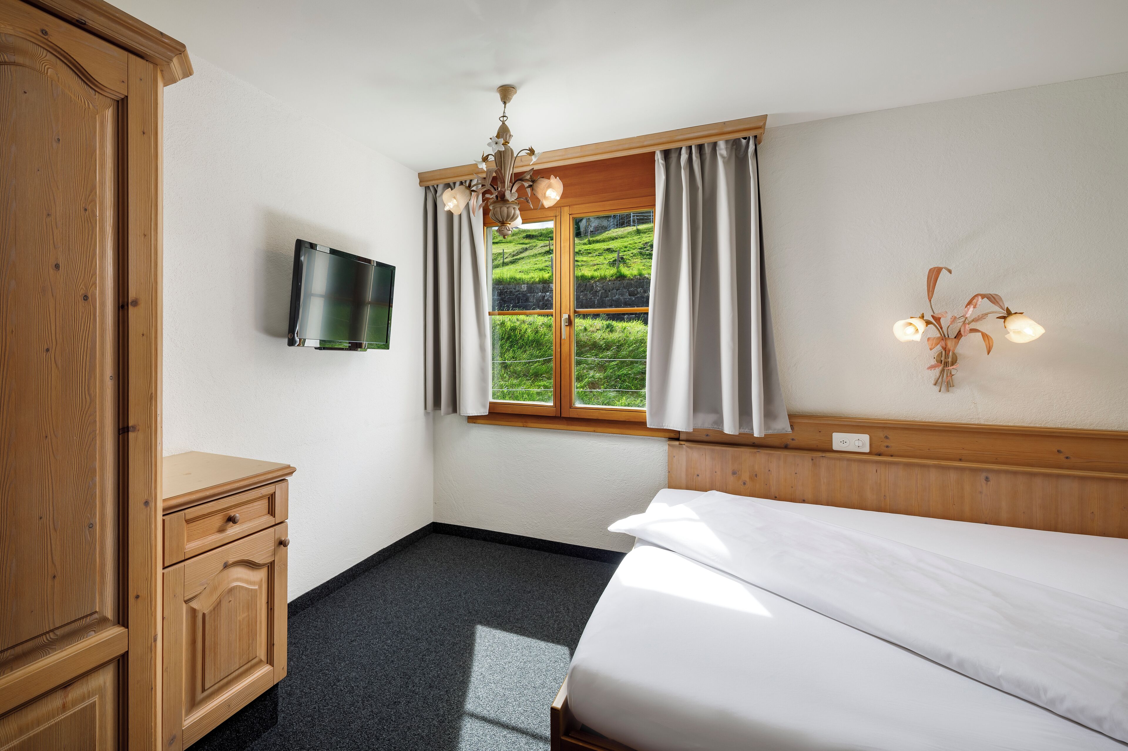 A bedroom with a bed, a wooden wardrobe, a television mounted on the wall, a window with curtains, and a wall-mounted light fixture.