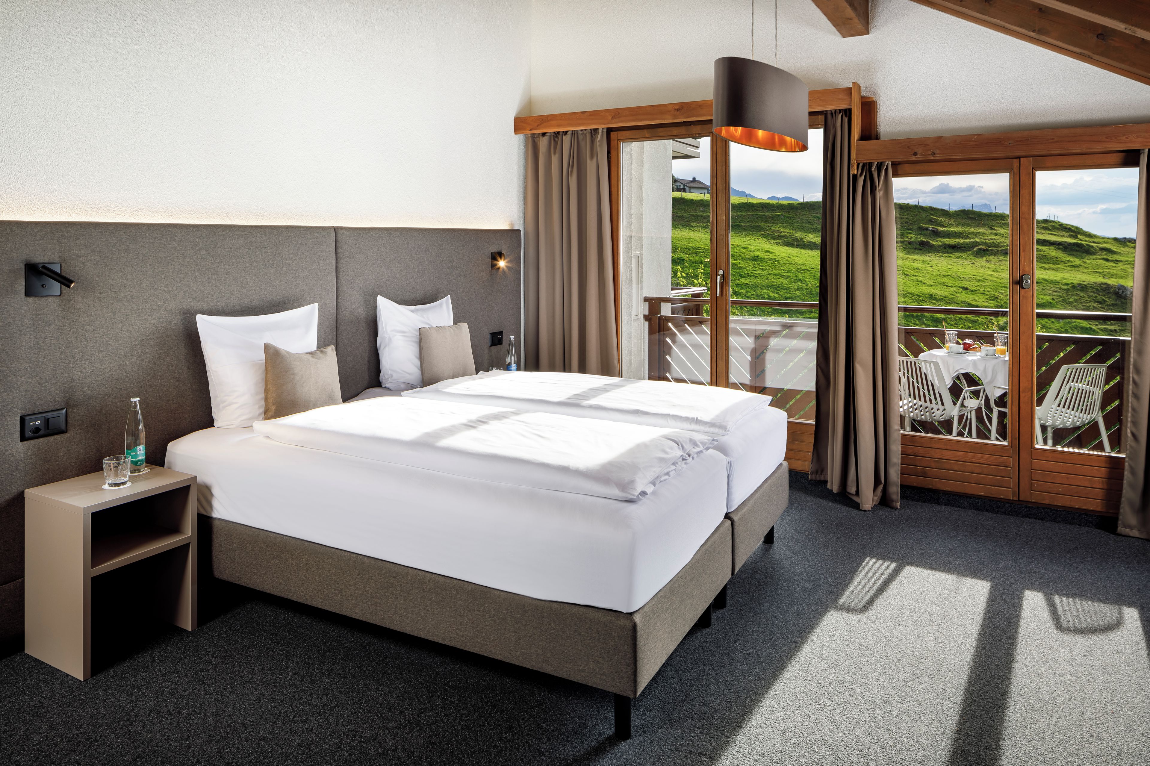 Habitación de hotel con una cama, mesitas de noche, una botella y un vaso. Se ve un balcón con mobiliario exterior.