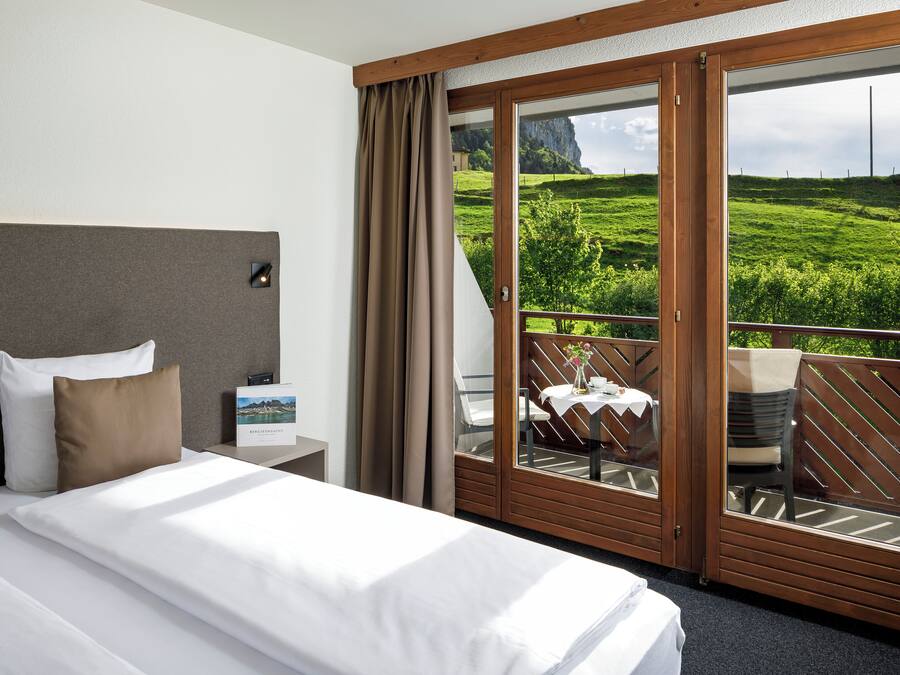 Habitación de hotel con cama, mesita de noche, vista a un balcón con mesa y sillas, y una vista panorámica al exterior.