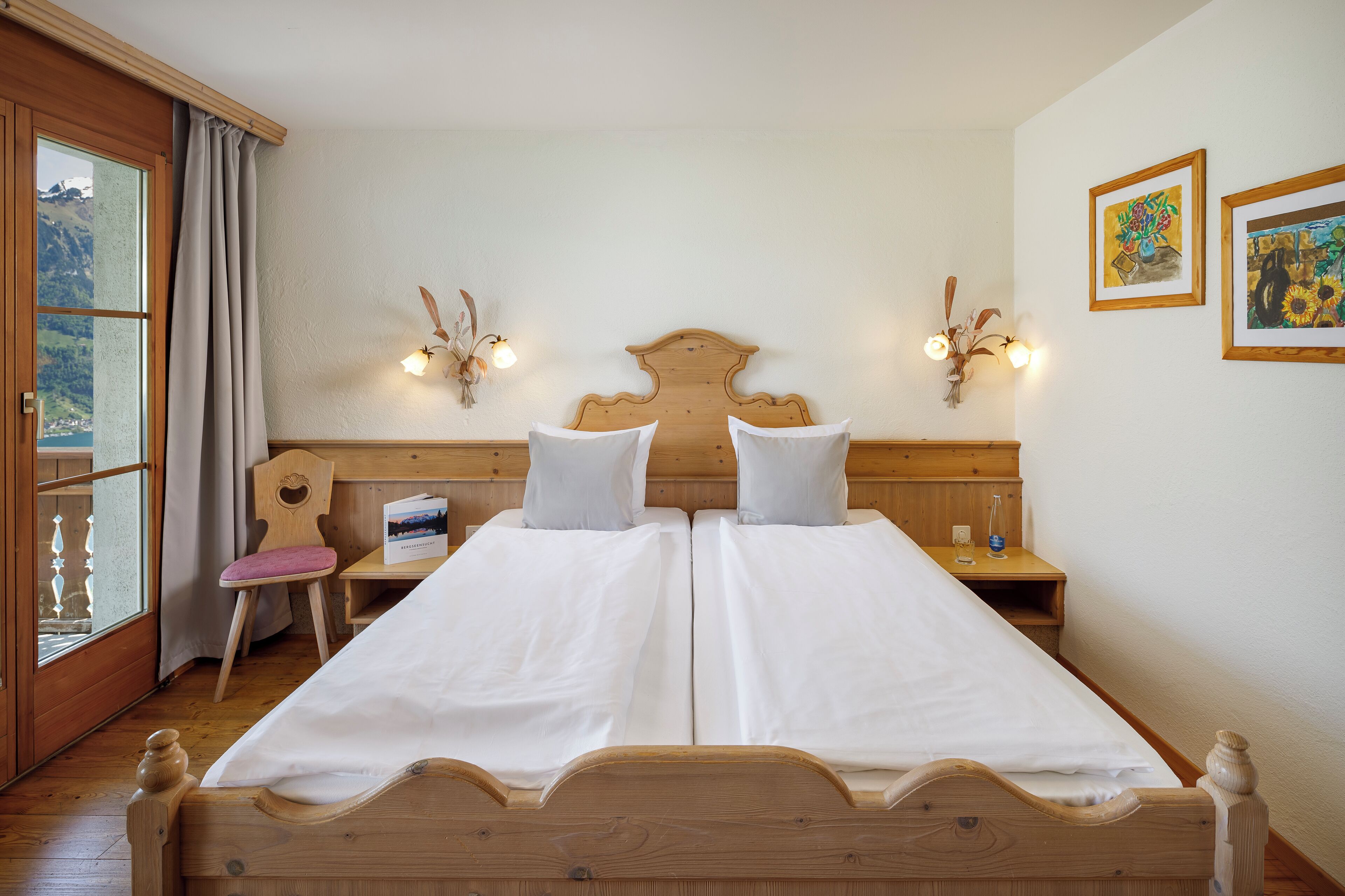 A double bed with white linens, wooden headboard, two bedside tables with lamps, a chair, and a window with curtains.