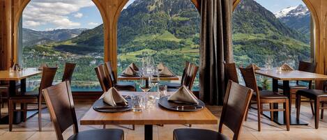 A dining area with tables set for four, large arched windows, and a mountainous landscape view.