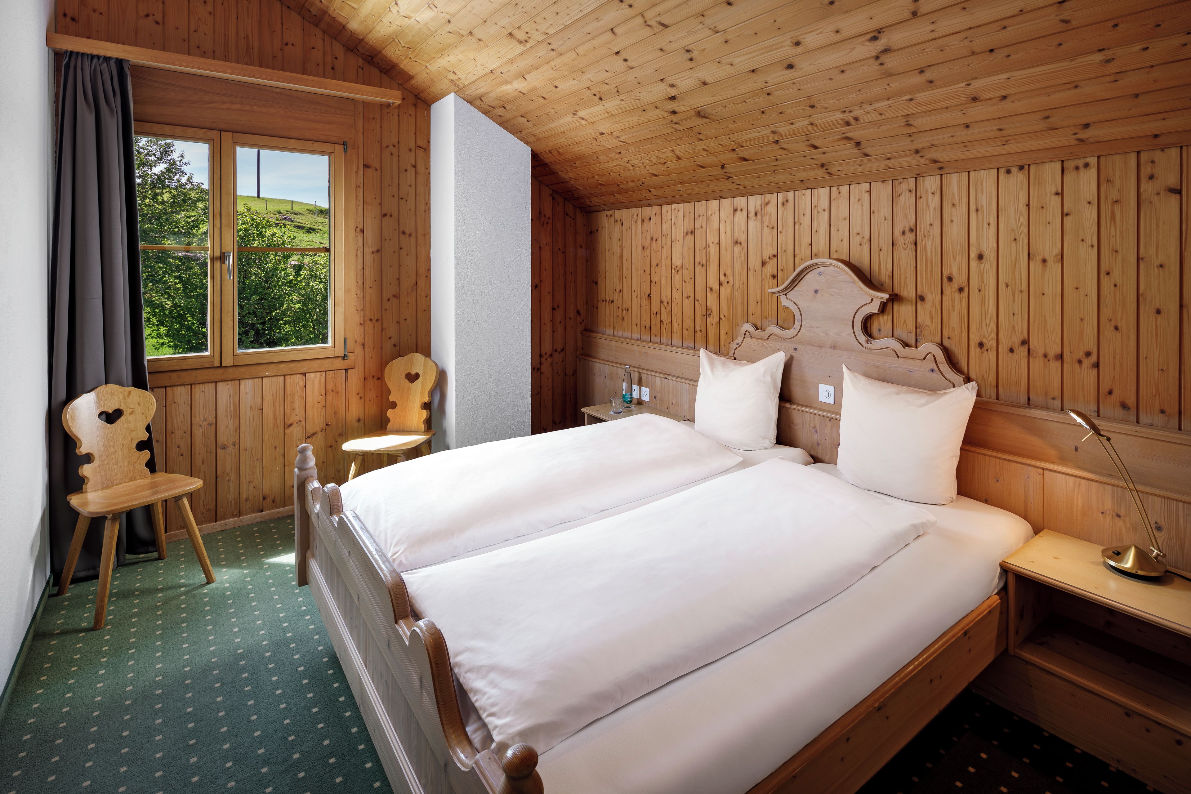 Habitación con paneles de madera, una cama doble, una silla, una mesita de noche y una ventana con vistas al follaje.