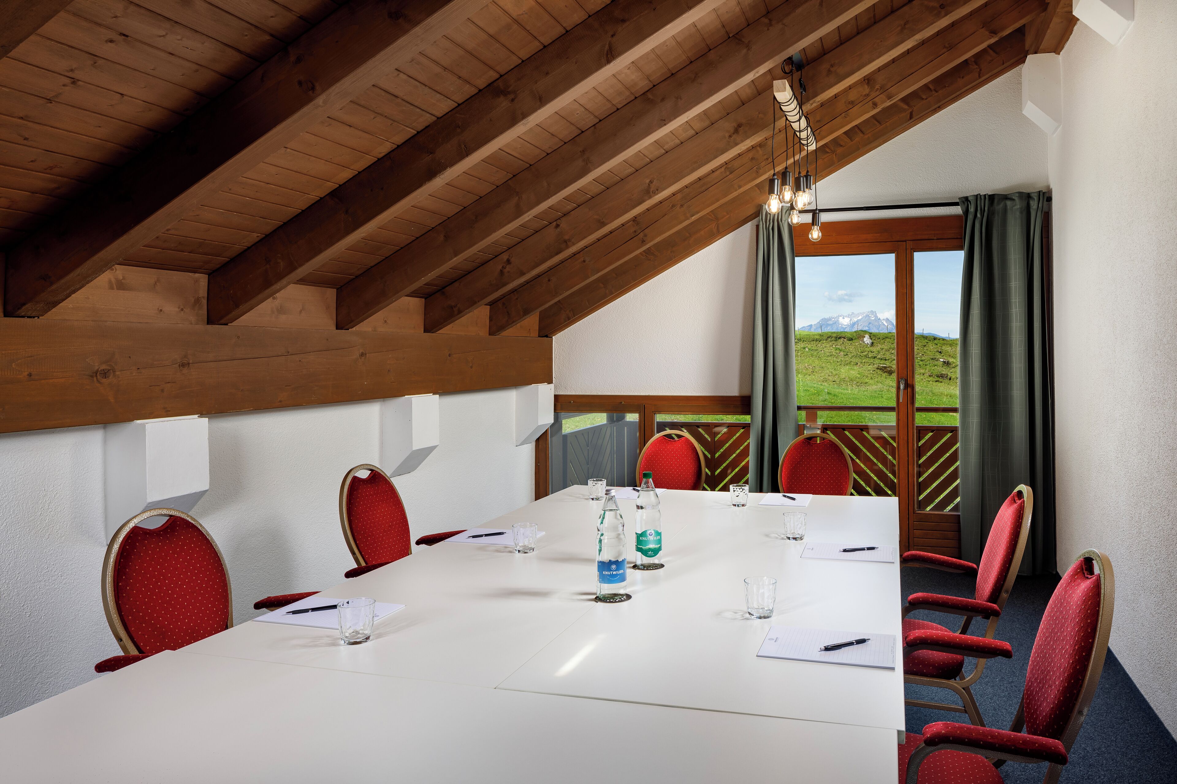Un salón de conferencias con una mesa blanca, sillas rojas y vistas a un paisaje verde a través de amplios ventanales.
