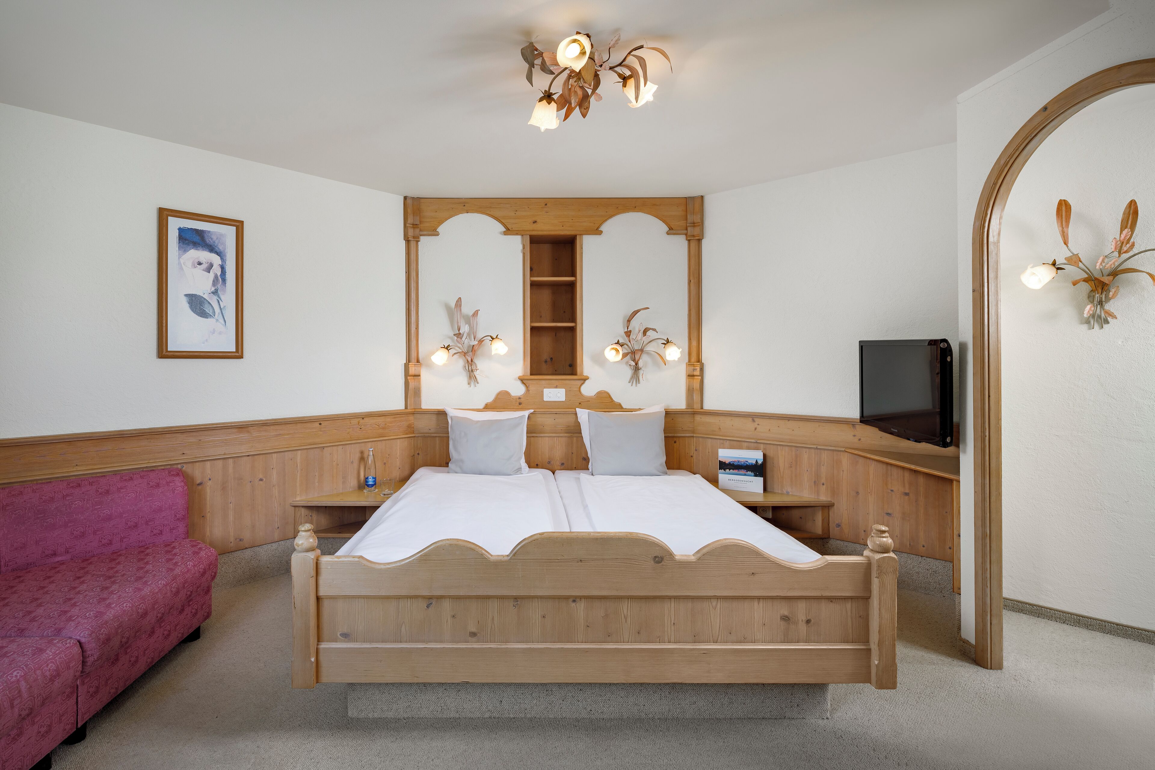 Ein Schlafzimmer mit einem hölzernen Bett, einem rosa Sofa, einem Nachttisch aus Holz, einem Spiegel und einer Deckenleuchte.