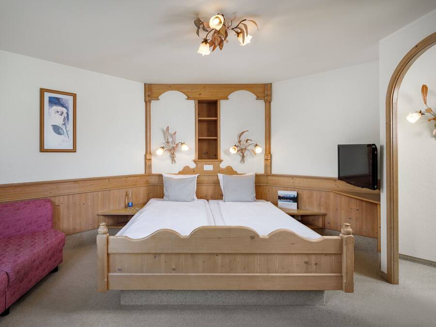 Un dormitorio con una cama de madera, un sofá rosa, una mesita de noche de madera, un espejo y una lámpara en el techo.