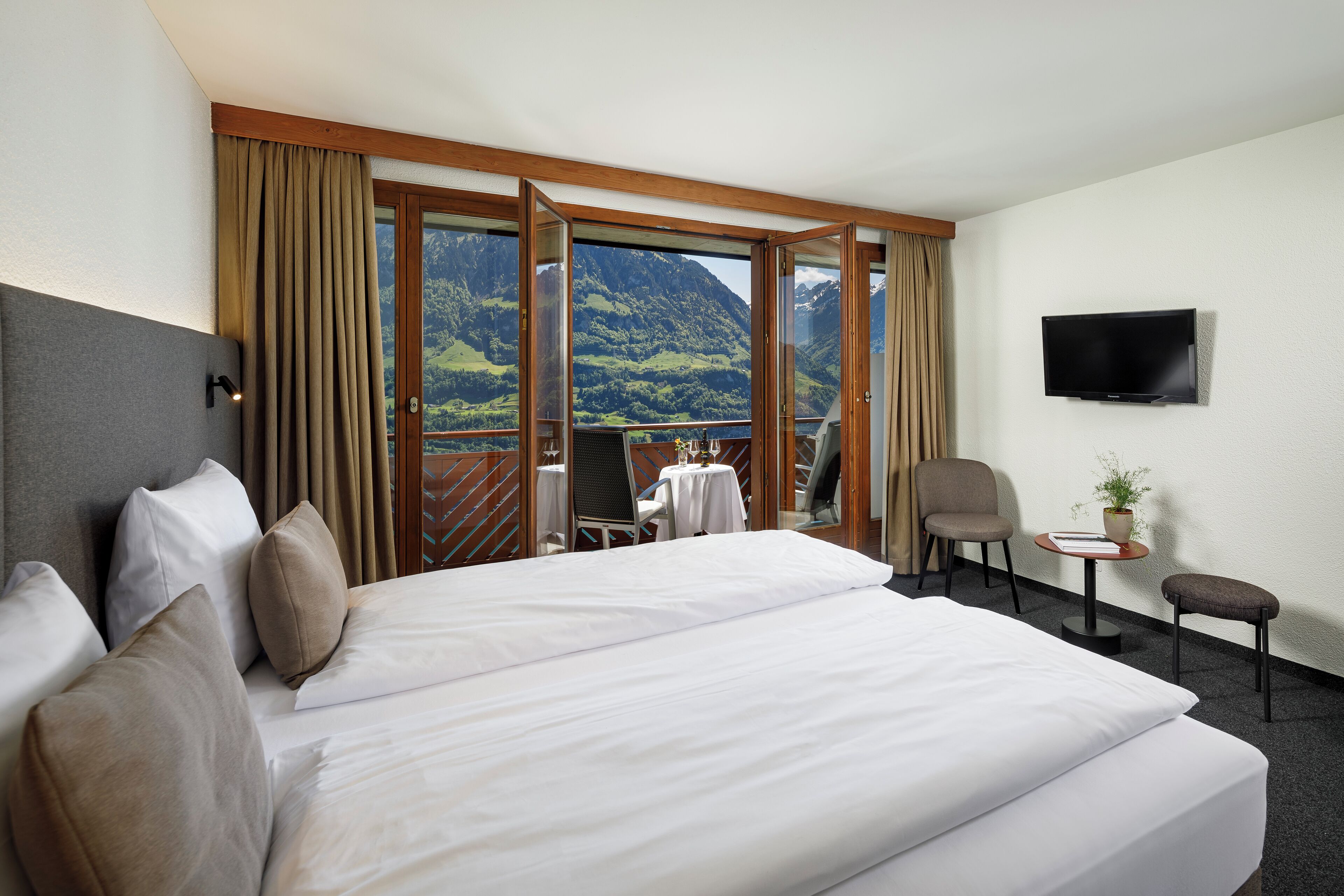 Habitación de hotel con una cama grande, televisor montado en la pared, balcón con vistas a la montaña y una mesita con una planta.
