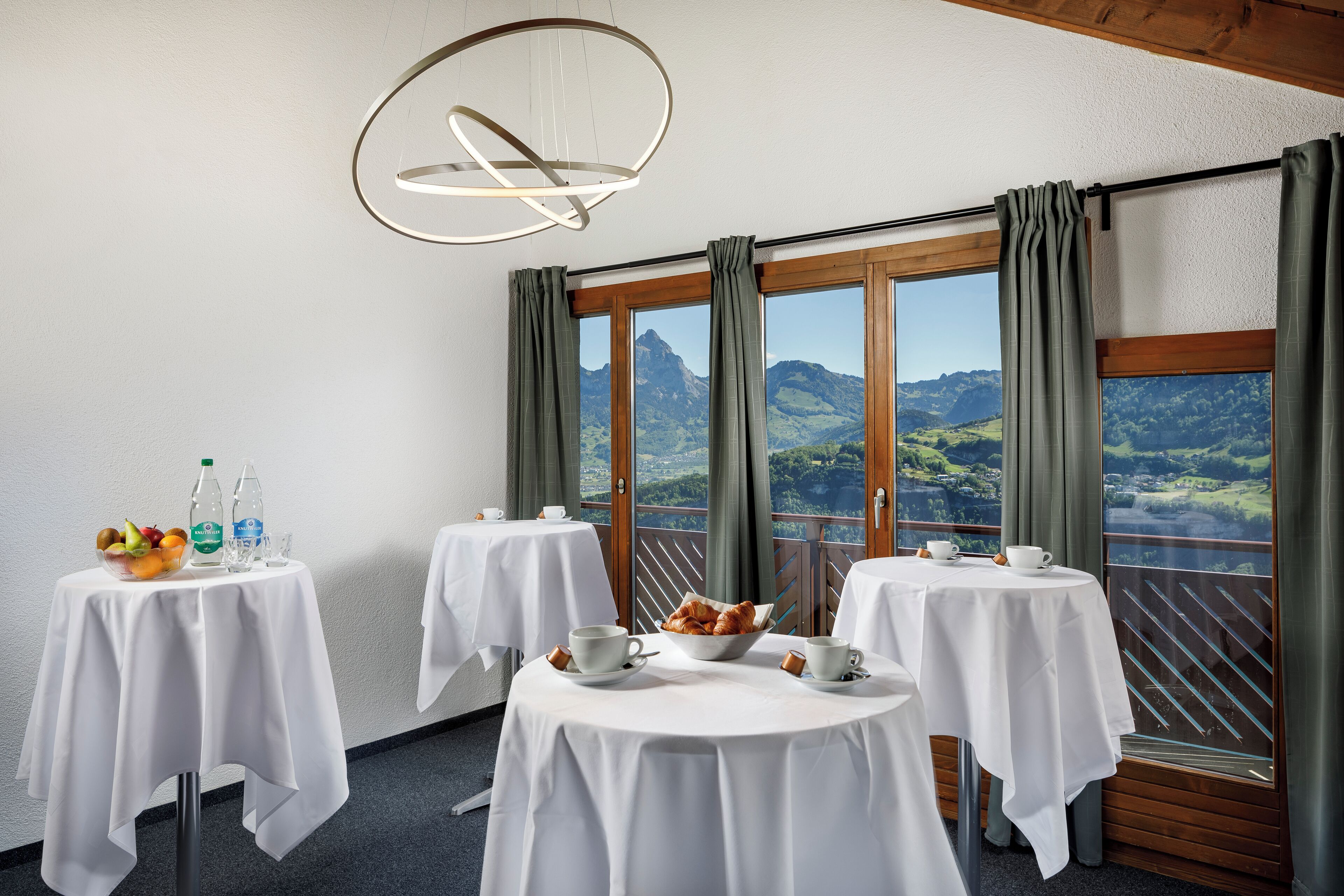 A dining area with three round tables, each with a white tablecloth and set with cups, saucers, and a bowl of pastries. There are two bottles of water and a bowl of fruit on the first table. The room has large windows with curtains, offering a view of green hills and a mountain in the distance.