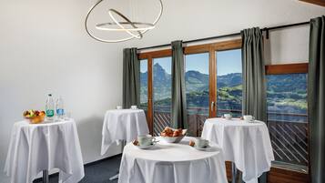 A dining area with three round tables, each with a white tablecloth and set with cups, saucers, and a bowl of pastries. There are two bottles of water and a bowl of fruit on the first table. The room has large windows with curtains, offering a view of green hills and a mountain in the distance.