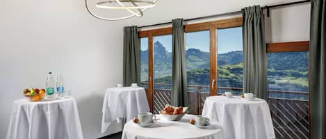 A dining area with three round tables, each with a white tablecloth and set with cups, saucers, and a bowl of pastries. There are two bottles of water and a bowl of fruit on the first table. The room has large windows with curtains, offering a view of green hills and a mountain in the distance.