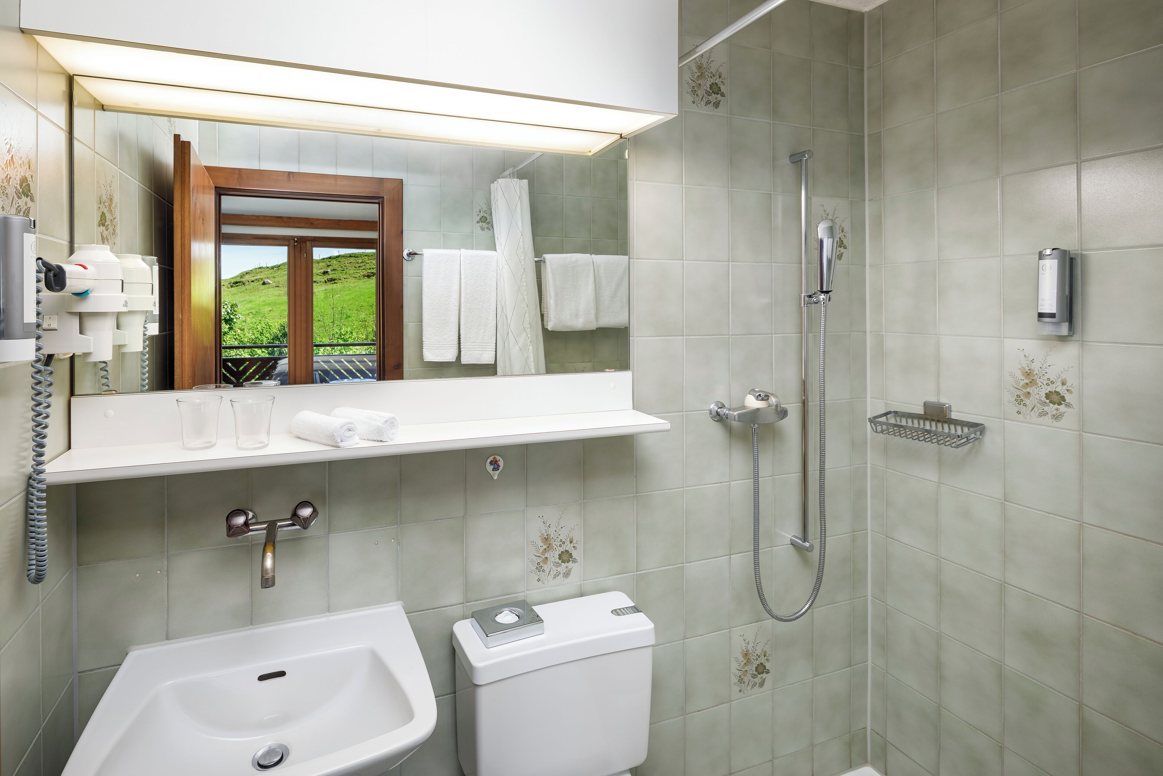Een badkamer met een wastafel, toilet en douche. Boven de wastafel hangt een spiegel, en een handdoekrek met handdoeken.