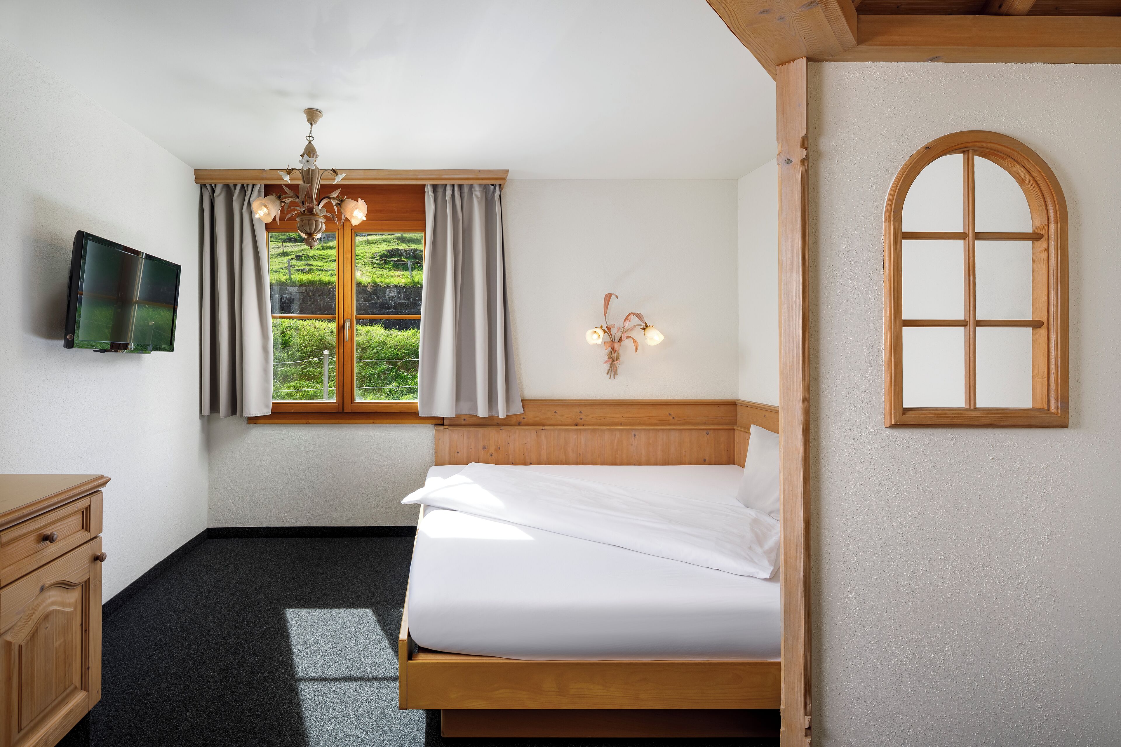 Ein Schlafzimmer mit Bett, Fernseher, Fenster mit Vorhängen, einer Chandelüster und einem verzierten, bogenförmigen Holzfenster.