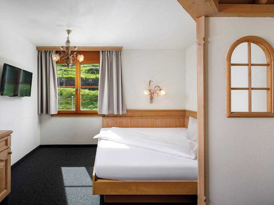 Ein Schlafzimmer mit Bett, Fernseher, Fenster mit Vorhängen, einer Chandelüster und einem verzierten, bogenförmigen Holzfenster.