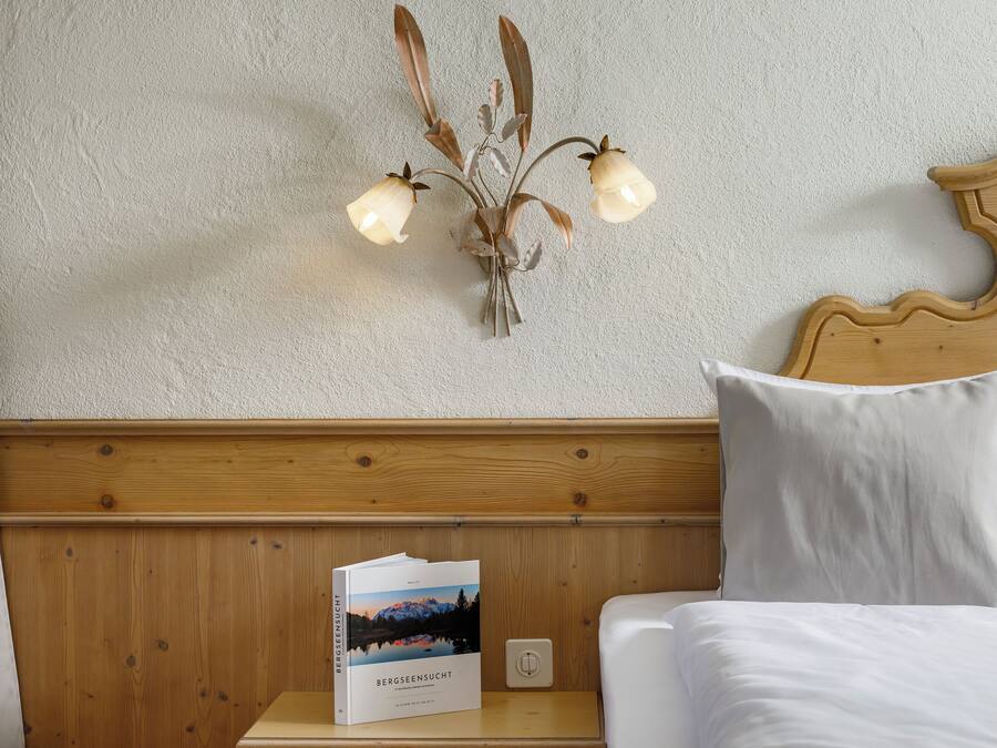 A neatly made bed with white linens, a wooden headboard, and a bedside table with a book titled "BERGEENSUCHT".