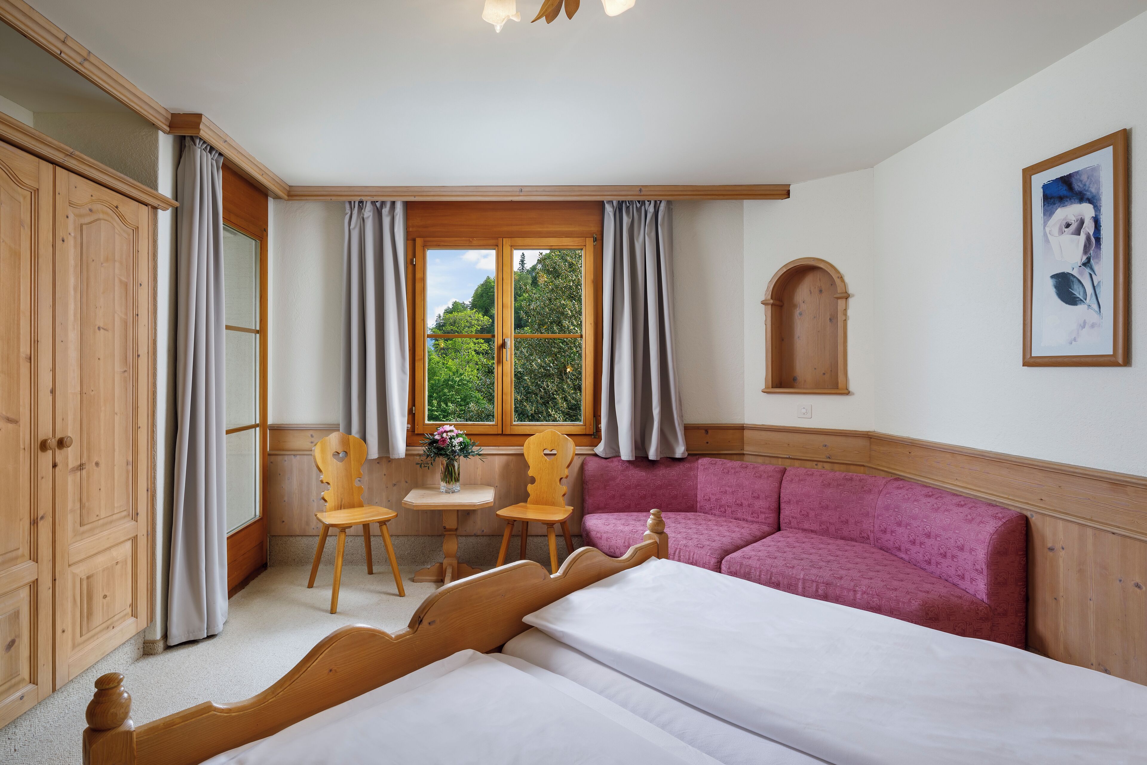 A room with a bed, a pink sofa, a wooden cabinet, a small table with chairs, and a window with curtains.
