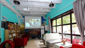 A room with a projector screen, a sofa, a table with a vase, and a window with curtains. - Meet int'l Youth Hostel Zhejiang Rd (Qingdao)