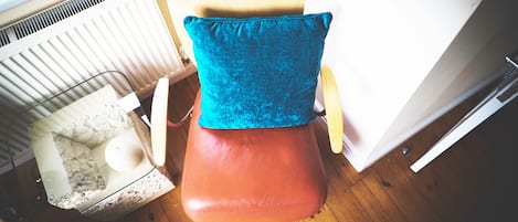 A corner of a room with a wooden floor, a radiator, a chair with a blue cushion, and a small pet bed.