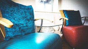 Twee stoelen met houten leuningen en blauwe kussens, geplaatst bij een raam met uitzicht op een stenen muur en een radiator.