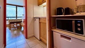 A compact kitchen with a microwave, coffee maker, and Siemens refrigerator. A wooden cabinet and a small table with a tablecloth are visible.