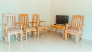 Een kamer met vijf houten stoelen en een tafel, een televisie op de tafel, en een vloer van witte tegels.